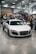 A sleek white sports car is displayed indoors at an automotive event. Several people are standing nearby, interacting and observing the cars. A tent with branding is in the background, along with other luxury vehicles parked in a spacious area with a high ceiling.