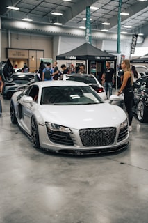 A sleek white sports car is displayed indoors at an automotive event. Several people are standing nearby, interacting and observing the cars. A tent with branding is in the background, along with other luxury vehicles parked in a spacious area with a high ceiling.