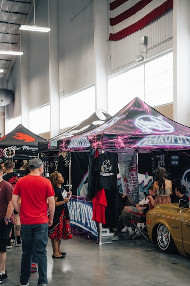 A lively indoor event scene with several people interacting near a series of colorful tent booths. Merchandise, such as t-shirts, is displayed prominently. The space has a high ceiling, and a large American flag is visible hanging in the background. Some attendees are engaged in conversation while others are browsing the goods. A vintage car is parked nearby, adding to the atmosphere.