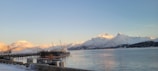 A large ship is docked at a pier against a backdrop of snow-covered mountains bathed in warm, golden light. The calm water reflects the mountain scenery, creating a tranquil atmosphere. The foreground shows a snow-covered dock area with some equipment and signage.