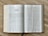 An open book displaying two pages with the title 'The Book of Tobit' on the left-hand page. The pages contain structured text in columns with headings and numbered sections. The book has a visible brown spine and rests on a wooden surface.