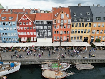 A row of colorful historic buildings lines a waterfront, with cafes and people gathered along the harbor. A traditional wooden sailing ship is moored in the water in front. The setting is lively and bustling with activity, suggesting a popular tourist or local spot.