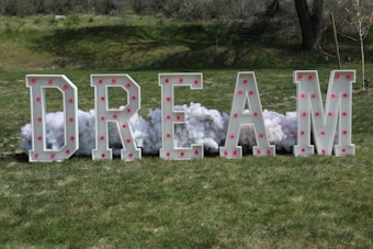 Large block letters spelling out the word 'DREAM' are standing on a grassy area. The letters are decorated with pink lights, and fluffy white material resembling clouds is arranged behind them.