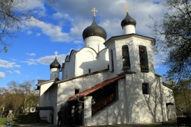 An ornate white building featuring classic Orthodox architectural elements, including large domes topped with crosses. The background shows a bright, partly cloudy sky framed by trees, with dappled light casting shadows on the building's facade.
