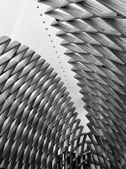 Intricate architectural structure with overlapping geometric patterns, creating a sense of movement and depth. The design is comprised of repeated wooden elements that form arch-like shapes, set against a contrasting white background with circular details.
