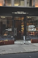 A storefront with large glass windows displaying the sign of a liquor-infused ice cream bar. The exterior features two planters with dry grass and modern wire-frame chairs. Inside, warm lighting illuminates a bar area, and a person is seated at a table writing or eating.