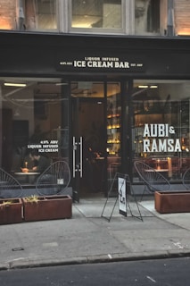 A storefront with large glass windows displaying the sign of a liquor-infused ice cream bar. The exterior features two planters with dry grass and modern wire-frame chairs. Inside, warm lighting illuminates a bar area, and a person is seated at a table writing or eating.