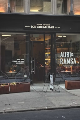 A storefront with large glass windows displaying the sign of a liquor-infused ice cream bar. The exterior features two planters with dry grass and modern wire-frame chairs. Inside, warm lighting illuminates a bar area, and a person is seated at a table writing or eating.