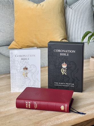 Three Bibles are displayed on a wooden surface. On the left, there is a white-covered booklet titled 'Coronation Bible' from The King’s Printer by Cambridge University Press, featuring a crowned gold emblem. In the middle is a black-covered booklet with the same title and presentation. The foreground showcases a red leather-bound Holy Bible, identified as the Authorized Version with a Cambridge publisher emblem.