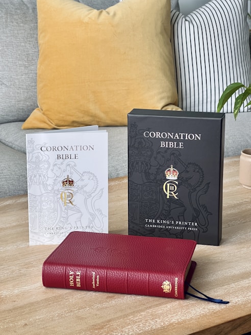 Three Bibles are displayed on a wooden surface. On the left, there is a white-covered booklet titled 'Coronation Bible' from The King’s Printer by Cambridge University Press, featuring a crowned gold emblem. In the middle is a black-covered booklet with the same title and presentation. The foreground showcases a red leather-bound Holy Bible, identified as the Authorized Version with a Cambridge publisher emblem.