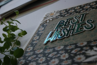 A greeting card prominently features the phrase 'Best Wishes' in shiny, reflective lettering set against a background with floral patterns. A sprig of green leaves lies beside the card, adding a touch of nature.
