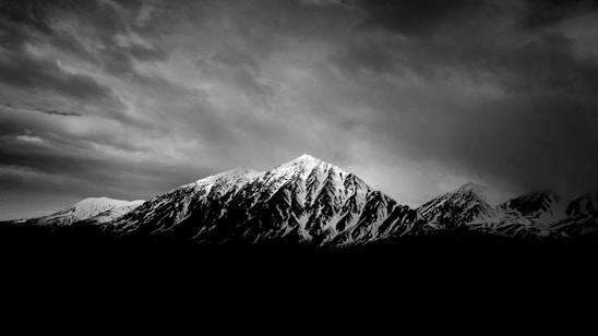 A black and white photograph of a mountain range with a prominent peak in the center. The mountains are covered in snow and the sky is cloudy, creating a moody atmosphere.