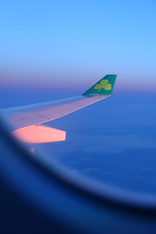 An airplane wing stretches out into a vibrant, colorful sky during either sunrise or sunset. The wingtip features a green logo with a clover emblem. The window frame of the airplane is visible in the foreground, giving a sense of perspective from within the cabin.