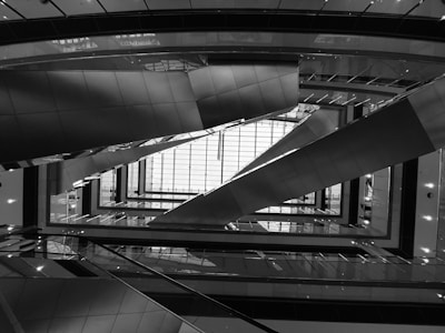 A complex architectural scene captured in black and white, focusing on intersecting lines and geometric patterns. The upward view highlights several escalators within a modern building, creating a sense of depth and perspective.