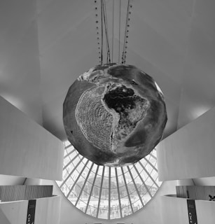 A large spherical object resembling Earth hangs suspended from the ceiling of a modern architectural space. The walls surrounding the sphere are smooth and angular, with large windows allowing natural light to illuminate the scene. The spherical object appears textured, displaying continents and oceans.