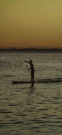 A person paddling a stand-up paddleboard on a calm river at sunset.