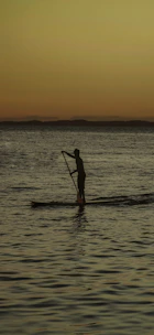 A person paddling a stand-up paddleboard on a calm river at sunset.