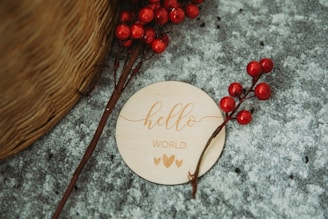 A wooden disc with the words 'hello world' and two hearts is placed on a textured gray surface. Nearby, red berry branches are arranged artistically. The edge of a woven basket is partially visible.