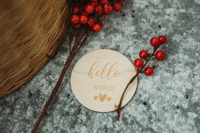 A wooden disc with the words 'hello world' and two hearts is placed on a textured gray surface. Nearby, red berry branches are arranged artistically. The edge of a woven basket is partially visible.