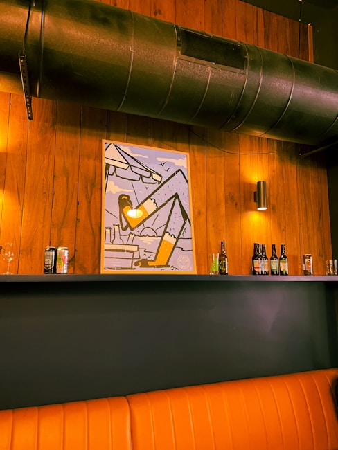 A warm-toned interior setting features a wooden wall with a large black ventilation pipe running across the top. Below the pipe, a colorful geometric poster is displayed. Several bottles and cans of drinks are neatly arranged on a shelf. The scene is softly lit by wall-mounted lamps, and a cushioned orange bench is partially visible at the bottom.