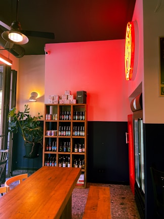 A cozy indoor space features a wooden table with benches, a wall-mounted shelf stocked with various bottles and boxes, and a refrigerator. A warm, ambient light from a neon sign bathes the room in a red glow, while natural light streams in from the left. A potted plant adds a touch of greenery to the setting, and the overall atmosphere is inviting and relaxed.
