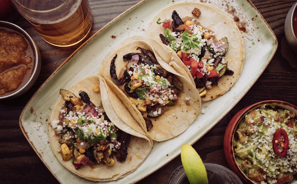 three tacos are sitting on a plate next to a glass of beer