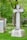 A peaceful cemetery scene with several large granite tombstones and ornate crosses surrounded by lush green grass and an abundance of leafy trees. Pink and white blooming flowers add a touch of color to the solemn yet serene setting.