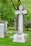 A peaceful cemetery scene with several large granite tombstones and ornate crosses surrounded by lush green grass and an abundance of leafy trees. Pink and white blooming flowers add a touch of color to the solemn yet serene setting.