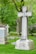 A peaceful cemetery scene with several large granite tombstones and ornate crosses surrounded by lush green grass and an abundance of leafy trees. Pink and white blooming flowers add a touch of color to the solemn yet serene setting.