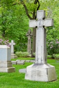 A peaceful cemetery scene with several large granite tombstones and ornate crosses surrounded by lush green grass and an abundance of leafy trees. Pink and white blooming flowers add a touch of color to the solemn yet serene setting.