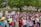 A large group of people gather outdoors holding various signs advocating for women's rights and equality. Many of the participants are wearing pink, and some have slogans on their shirts. The background features a public park with trees and a building, and the mood is one of unity and activism.