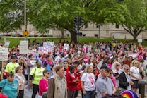 A large group of people gather outdoors holding various signs advocating for women's rights and equality. Many of the participants are wearing pink, and some have slogans on their shirts. The background features a public park with trees and a building, and the mood is one of unity and activism.