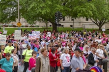 A large group of people gather outdoors holding various signs advocating for women's rights and equality. Many of the participants are wearing pink, and some have slogans on their shirts. The background features a public park with trees and a building, and the mood is one of unity and activism.