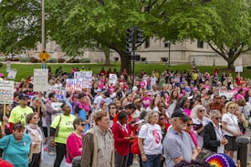 A large group of people gather outdoors holding various signs advocating for women's rights and equality. Many of the participants are wearing pink, and some have slogans on their shirts. The background features a public park with trees and a building, and the mood is one of unity and activism.
