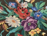 An intricate embroidery of colorful flowers, including red, purple, white, blue, and yellow blossoms, set against dark green foliage. The stitches are tightly packed, creating a detailed and vibrant tapestry.
