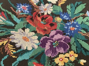 An intricate embroidery of colorful flowers, including red, purple, white, blue, and yellow blossoms, set against dark green foliage. The stitches are tightly packed, creating a detailed and vibrant tapestry.