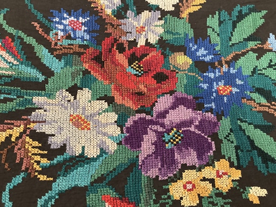 An intricate embroidery of colorful flowers, including red, purple, white, blue, and yellow blossoms, set against dark green foliage. The stitches are tightly packed, creating a detailed and vibrant tapestry.