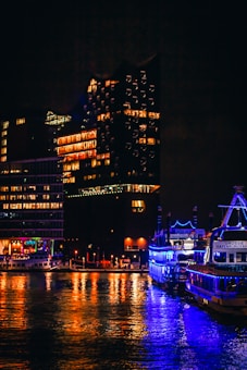 A nighttime cityscape by the water showcasing a brightly lit cruise ship with a vivid blue light display. The reflections of the ship and nearby buildings create colorful patterns on the water's surface. The background features modern buildings illuminated by warm lights, giving the scene a vibrant urban feel.