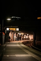 A dimly lit train station platform during nighttime with a small group of people huddled together near the edge. The platform curves around railroad tracks, and digital signs display information above. A solitary person stands to the left, illuminated by a streetlight.