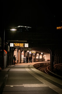 A dimly lit train station platform during nighttime with a small group of people huddled together near the edge. The platform curves around railroad tracks, and digital signs display information above. A solitary person stands to the left, illuminated by a streetlight.