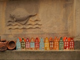 A row of colorful ceramic miniature houses is displayed against a textured stone wall. Above the houses, a carved design of a fish swimming above waves is prominently featured. The ceramics are painted in vibrant colors including orange, blue, green, and red.
