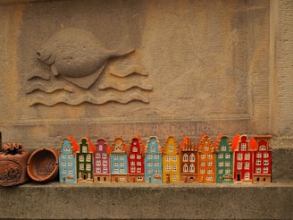 A row of colorful ceramic miniature houses is displayed against a textured stone wall. Above the houses, a carved design of a fish swimming above waves is prominently featured. The ceramics are painted in vibrant colors including orange, blue, green, and red.