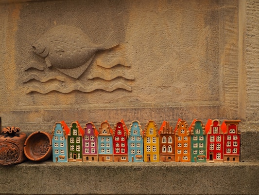 A row of colorful ceramic miniature houses is displayed against a textured stone wall. Above the houses, a carved design of a fish swimming above waves is prominently featured. The ceramics are painted in vibrant colors including orange, blue, green, and red.