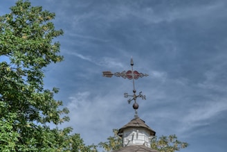 A weather vane sits atop a small wooden structure against a partly cloudy sky. The vane has a decorative arrow with cardinal directions marked.