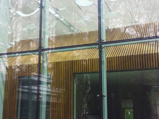The image depicts a modern architectural structure featuring large glass panels with metal frames. The interior visible through the glass includes walls with vertical wooden paneling. The reflections on the glass suggest surrounding trees or foliage, which contribute to a blend of natural and man-made elements.