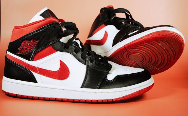 A pair of black, white, and red sneakers is positioned against a light orange background. The sneakers feature a high-top design with a prominent red swoosh logo on the side. The soles are red with black treads, and the upper part consists of a combination of leather materials with black laces.