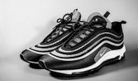 A pair of stylish sneakers with a modern design featuring a black and white color scheme. The shoes have a wave-like pattern on the sides, detailed lacing, and a sleek silhouette. The background is plain, emphasizing the footwear.
