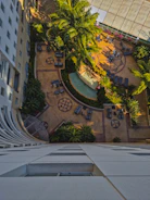 Aerial view of a courtyard with geometric-shaped tiles and lush green palm trees. The space includes several seating arrangements with tables and chairs, surrounded by dense greenery. A multi-story building with numerous windows frames one side of the courtyard.