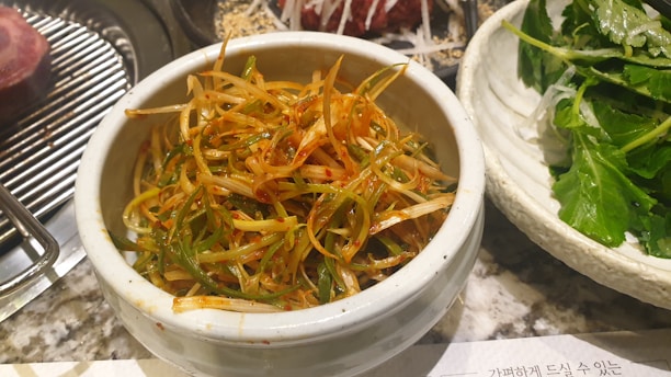 A bowl filled with spicy marinated green onion salad, surrounded by plates of leafy greens and grilled meat on the side.