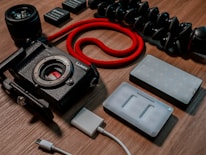 A collection of photography equipment is arranged on a wooden surface, including a black digital camera body without a lens, a red coiled cable, camera batteries, a flexible tripod, a camera lens, a USB cable, and LED light panels.